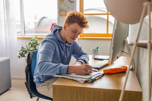 Teen in a blue hoodie doing school work at a computer and desk