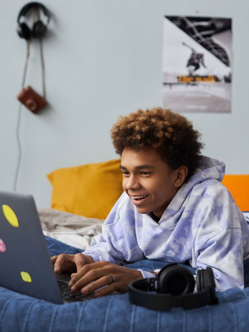 Young male in a hoodie uses a laptop while laying in bed