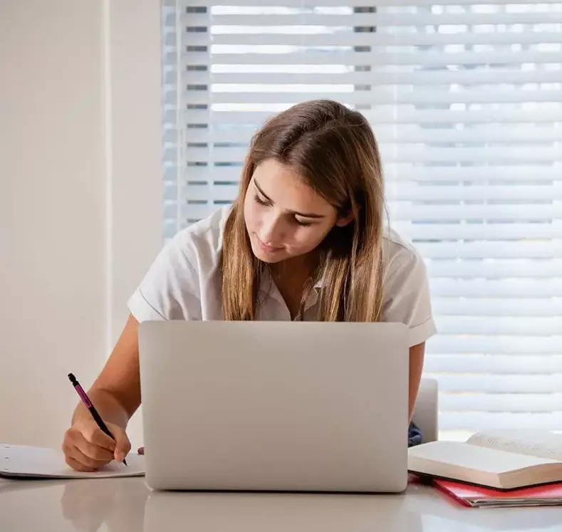 student writing in notebook while using laptop