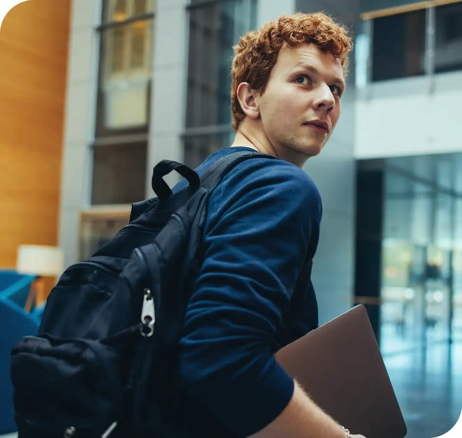 Student with laptop and backpack looking over his shoulder as he enters the building