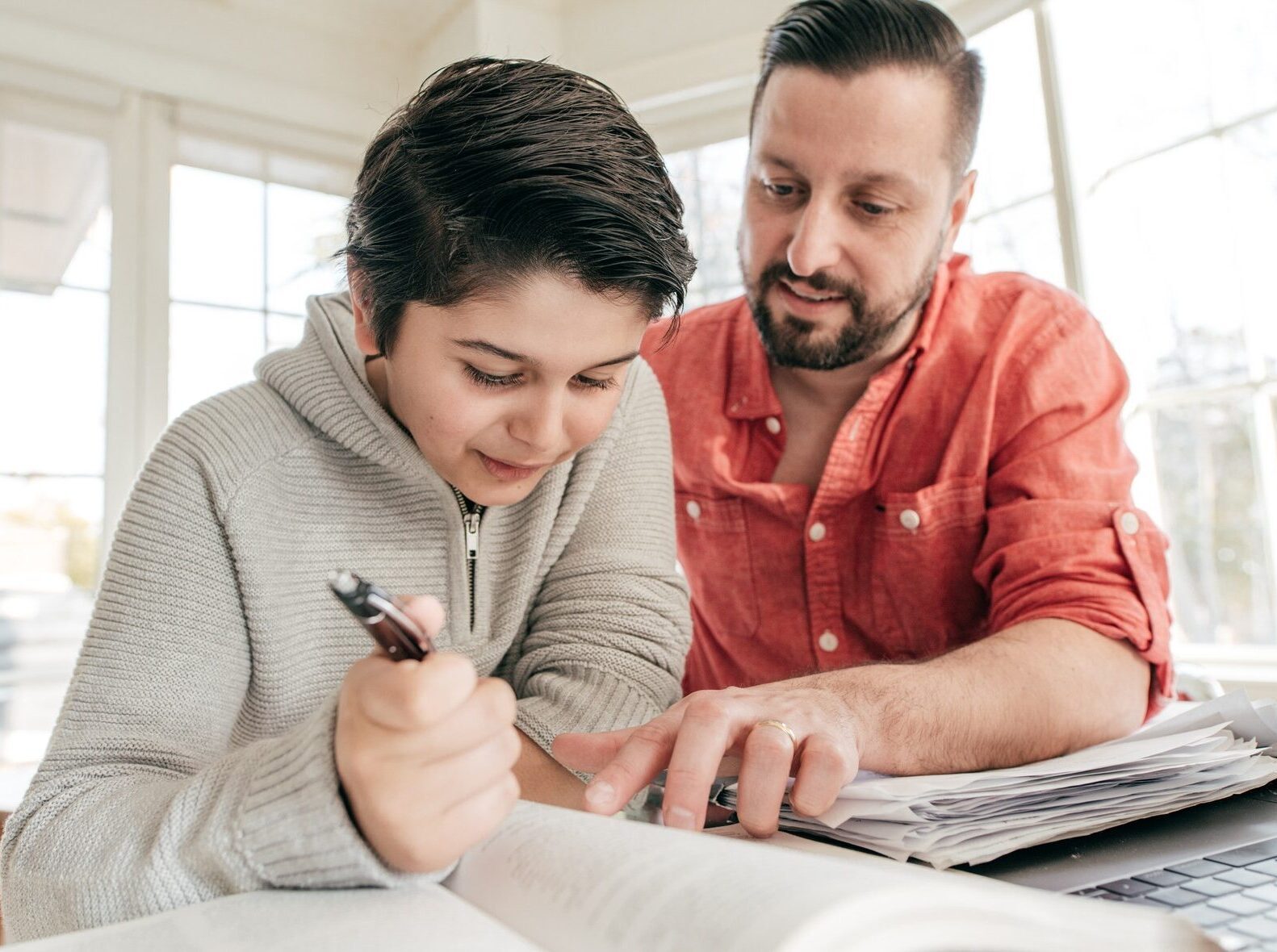 Boy with brown hair wearing a grey hoodie doing homework with a man in a red shirt