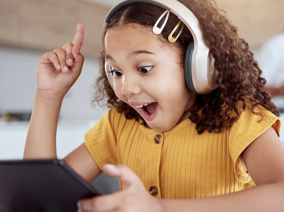 Young girl with brown curly hair wearing headphones and a yellow sweater exclaiming while using an ipad